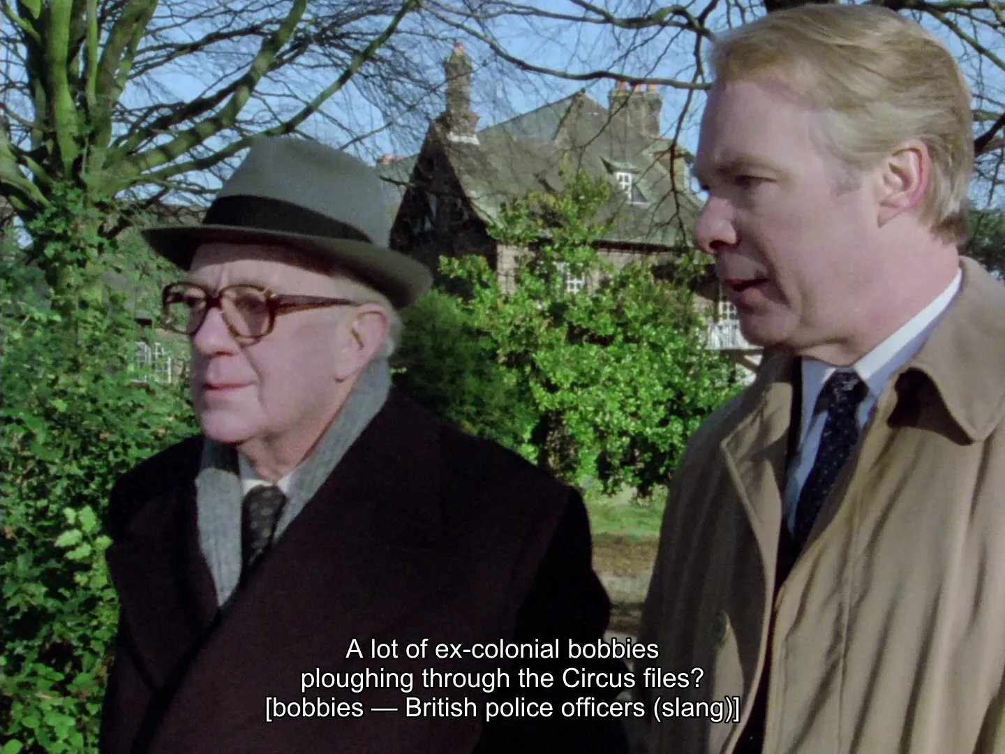 The same frame as the bobbies shot above, now on the English + Explainers track: the dialogue "A lot of ex-colonial bobbies ploughing through the Circus files?" is subtitled with "[bobbies — British police officers (slang)]" in brackets beneath it