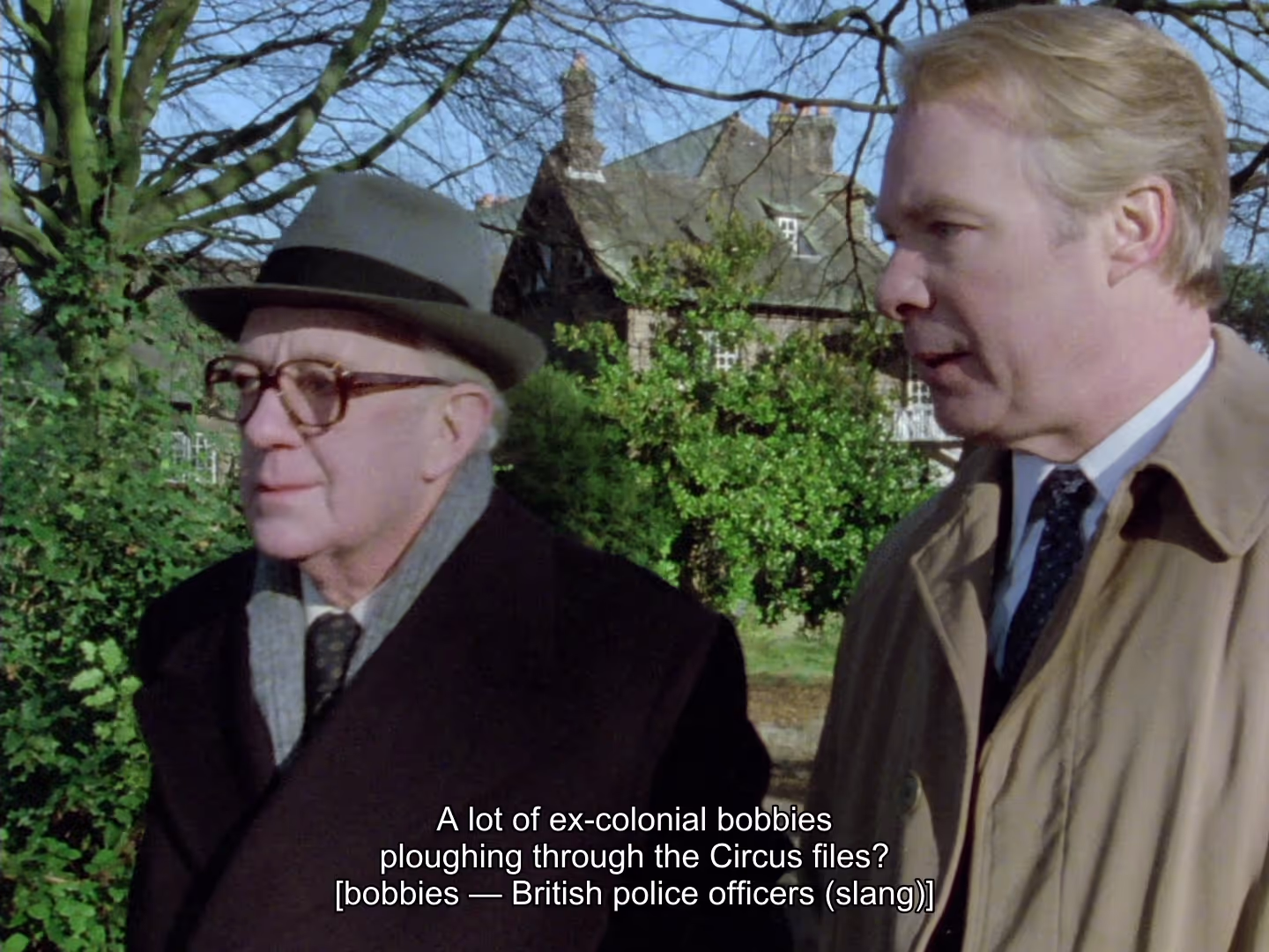 The same frame as the bobbies shot above, now on the English + Explainers track: the dialogue “A lot of ex-colonial bobbies ploughing through the Circus files?” is subtitled with “[bobbies — British police officers (slang)]” in brackets beneath it